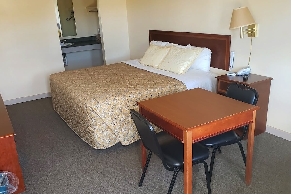 A hotel room with a bed, a desk with a chair, a lamp, and a small kitchenette.