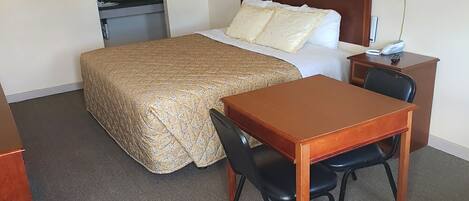 A hotel room with a bed, a desk with a chair, a lamp, and a small kitchenette.