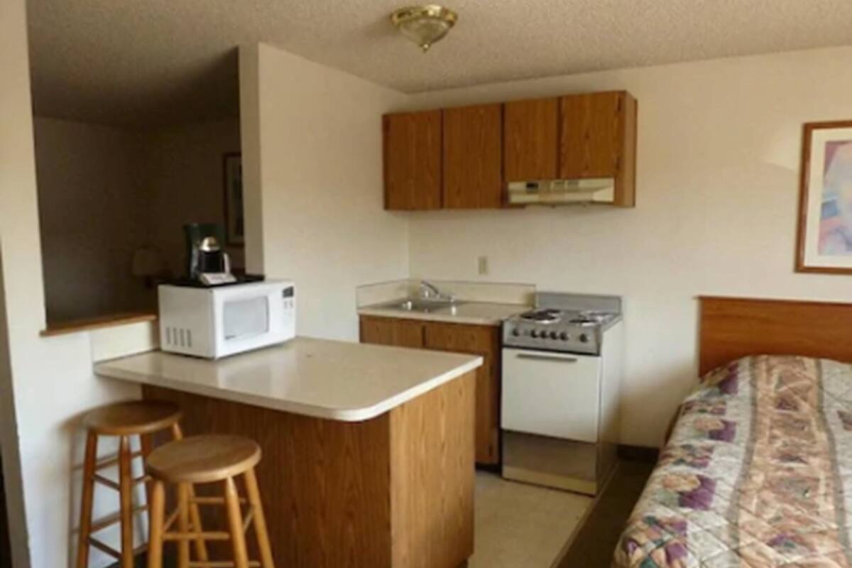 A compact kitchen with a microwave, stove, and wooden cabinets. There are two stools and a bed with a quilted blanket.