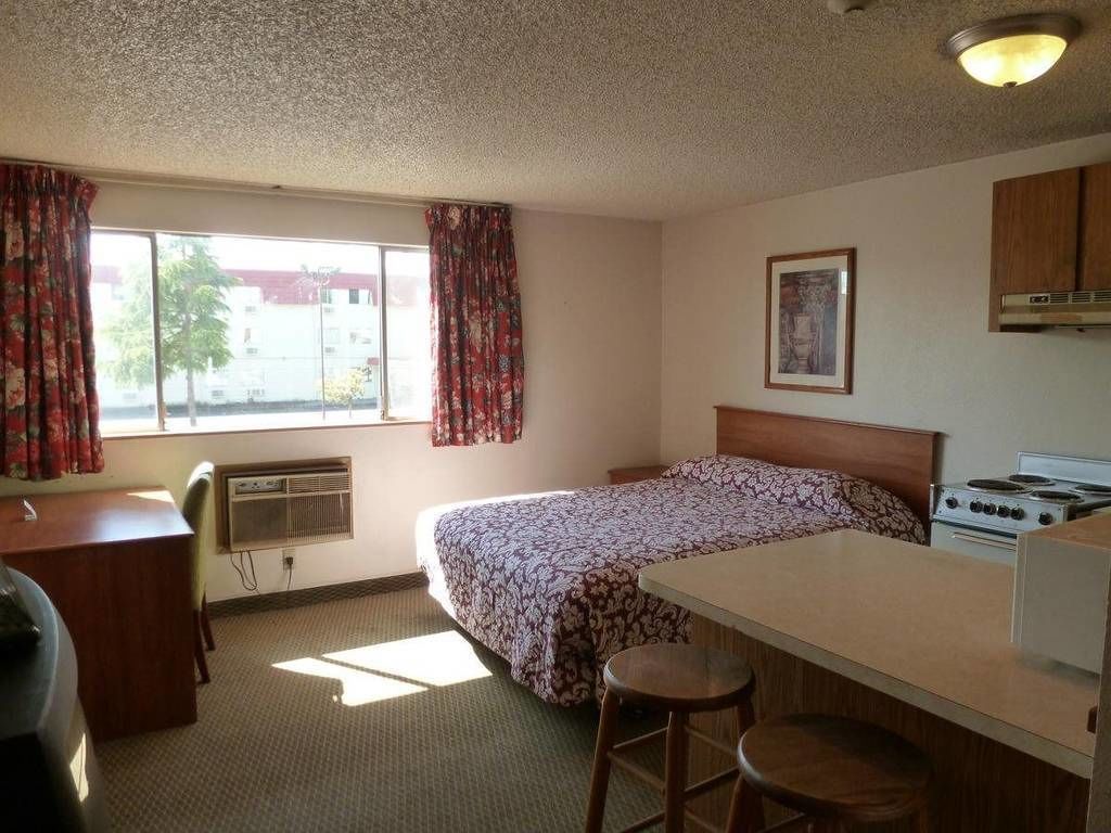 A small, compact room with a bed, a desk, a kitchenette, and a window with curtains.