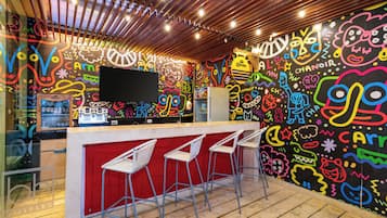 Una zona de bar con techo de madera, murales coloridos en las paredes, un televisor de pantalla plana y taburetes blancos.