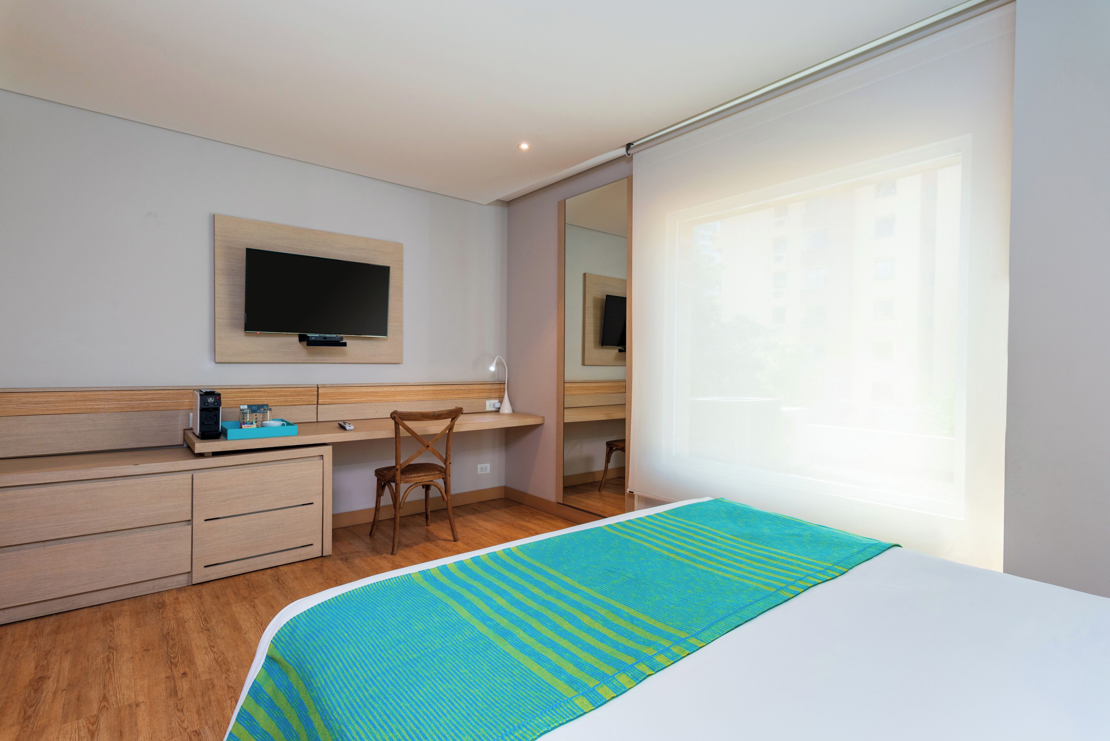 A modern hotel room with a bed, a desk, a chair, a TV mounted on the wall, and a window with curtains.