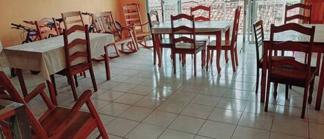 A dining area with multiple tables and chairs, large windows with a view of greenery, and a tiled floor.