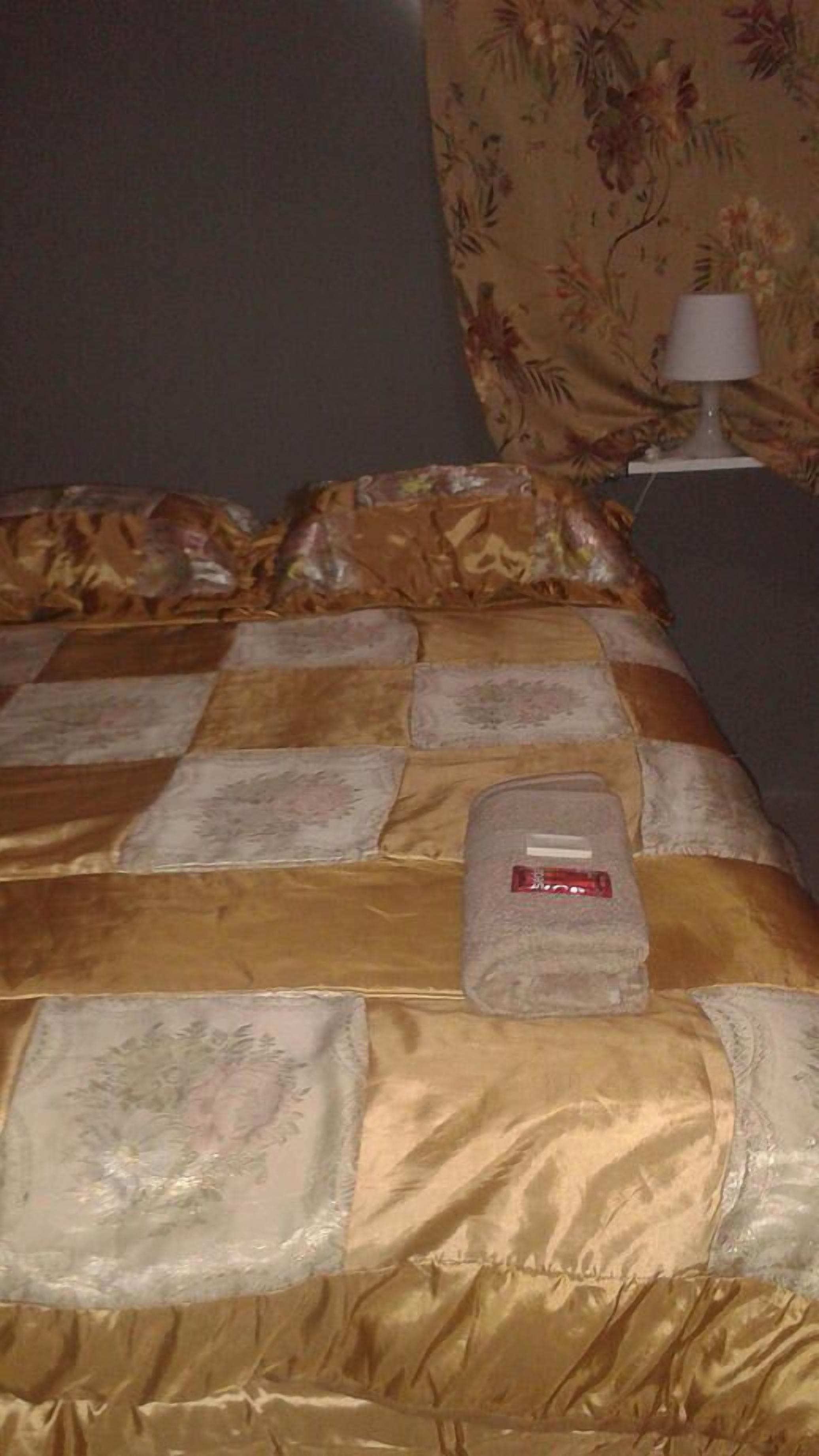 Una cama bien hecha con edredón estampado con motivos florales, una toalla doblada y una lámpara de noche en una mesita.