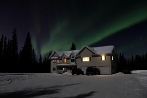 A house with a garage and a driveway, surrounded by snow and trees, under a night sky with the Northern Lights.