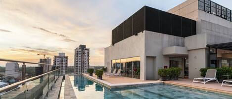 Piscina en la azotea con sillones de descanso, barandilla de vidrio y vistas de edificios circundantes.