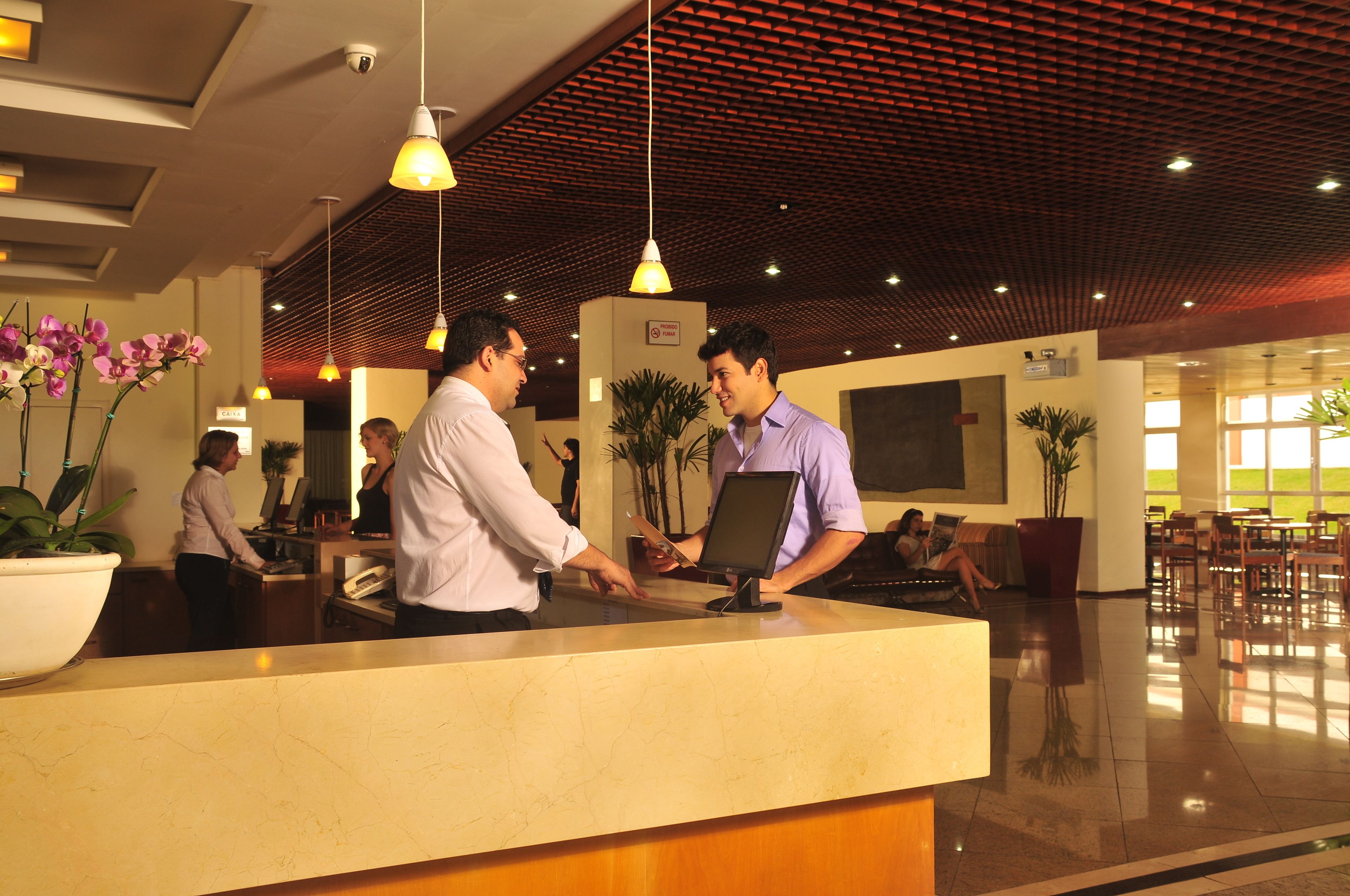 Uma área de recepção de hotel com balcão de mármore, iluminação pendente, plantas em vasos e um espaço de estar com mesas e cadeiras.