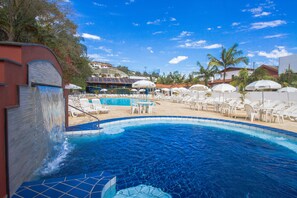 A swimming pool area with lounge chairs and umbrellas, surrounded by palm trees and a building in the background. - Hotel Majestic (Águas de Lindóia)