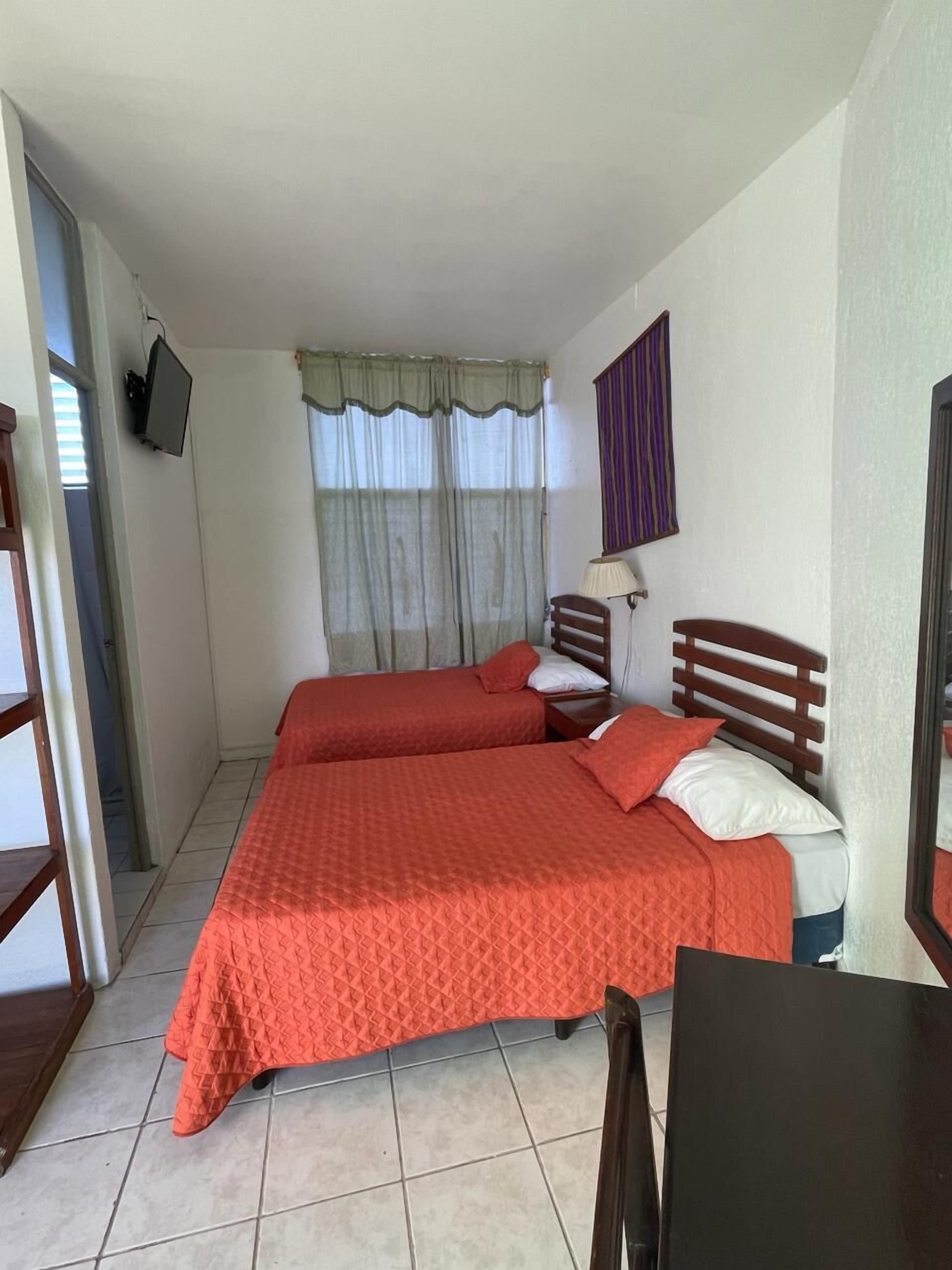 A room with two beds, a TV mounted on the wall, a window with curtains, and a small table.