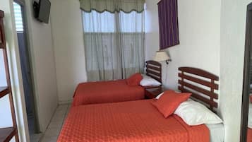 A room with two beds, a TV mounted on the wall, a window with curtains, and a small table.