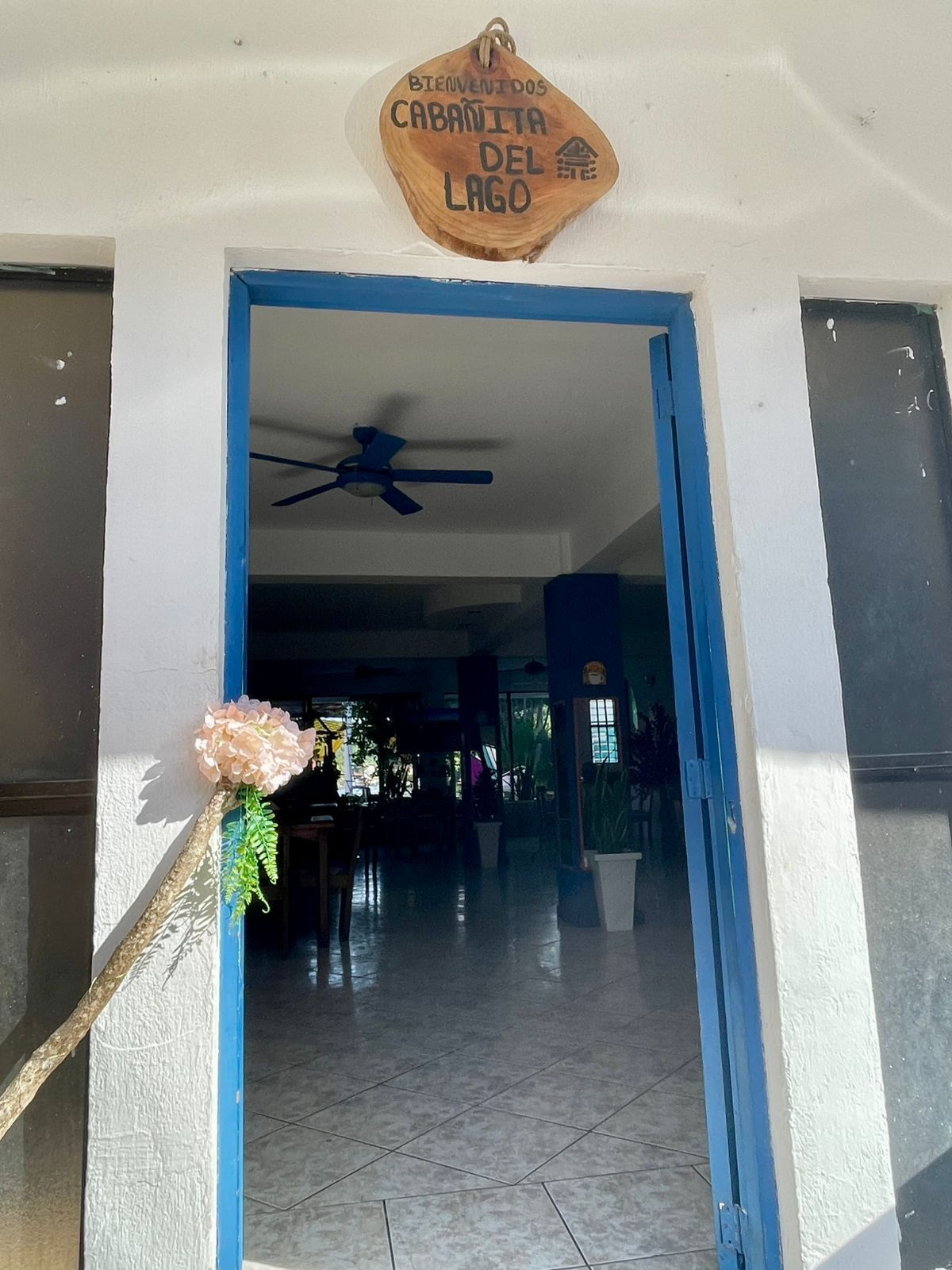 A wooden welcome sign with "BIENVENIDOS CABANITA DEL LAGO" text, a blue door frame, and a ceiling fan.