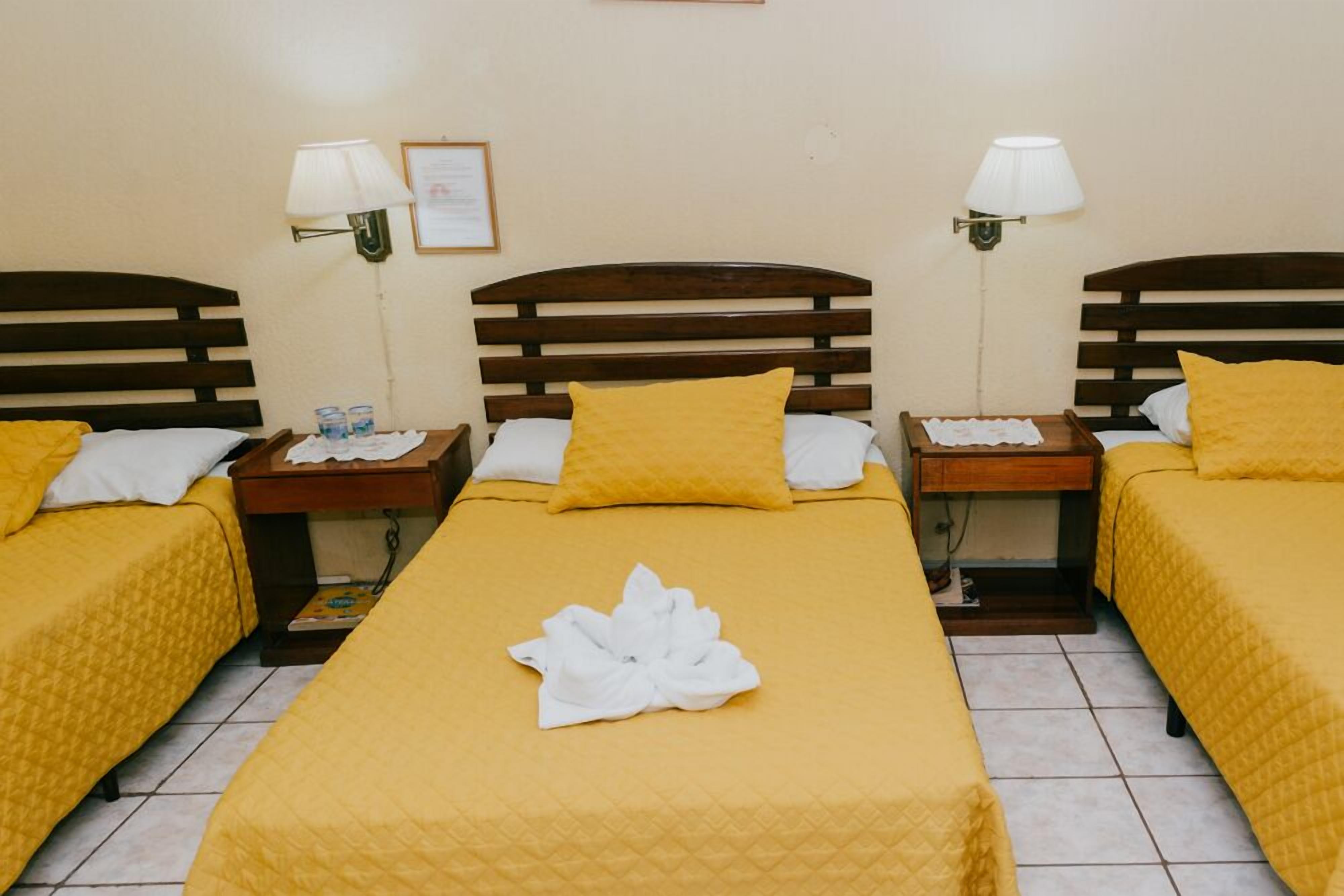 Habitación de hotel con tres camas, cada una con una colcha amarilla y almohadas blancas. Hay dos lámparas de pared a cada lado de la habitación.