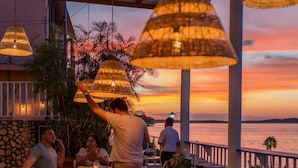 Zona de comedor al aire libre con mesas y sillas, con vista a un cuerpo de agua durante el atardecer.