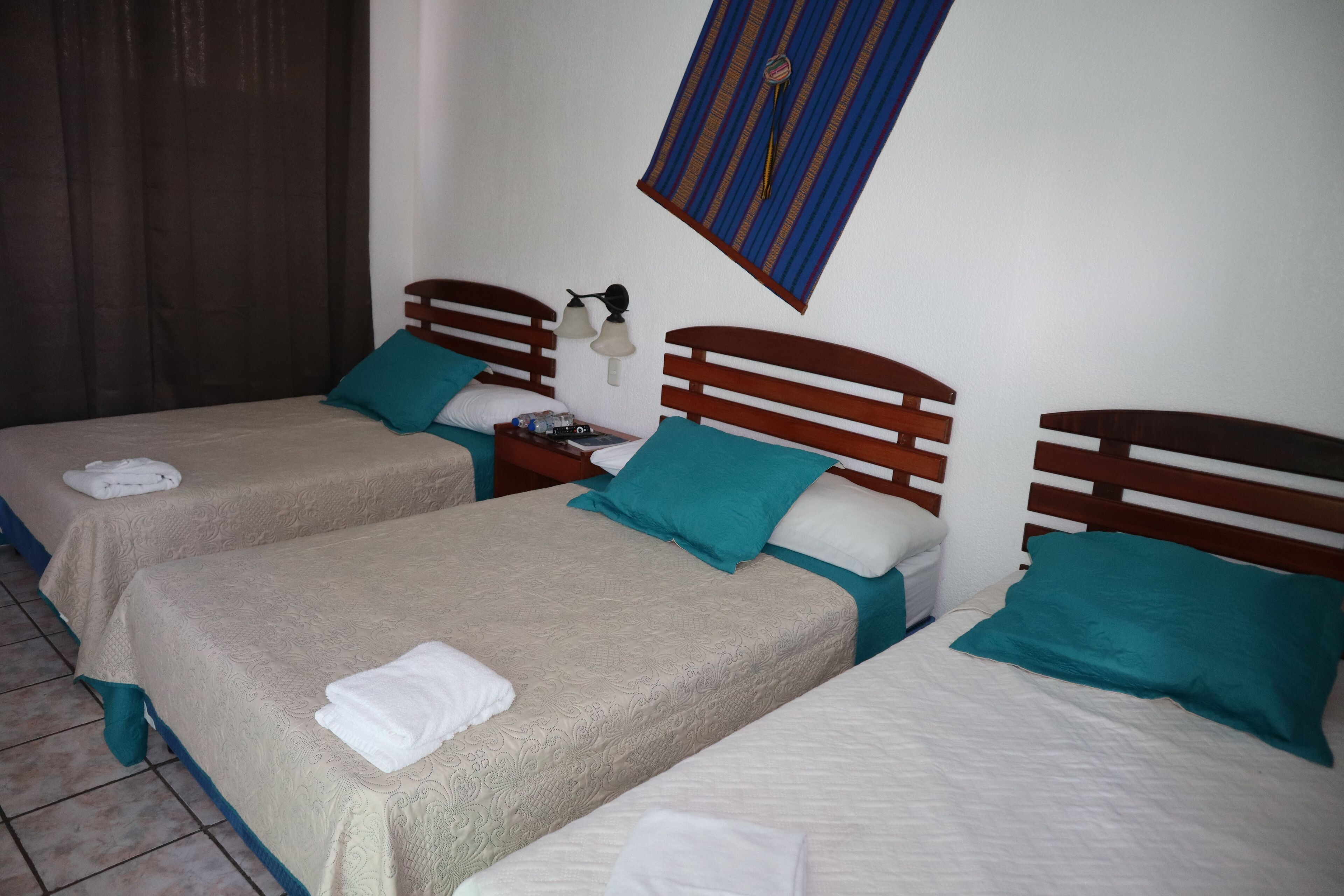 Habitación de hotel con dos camas, cada una con cabecero de madera y almohadas color turquesa. Hay una mesita de noche con lámpara entre las camas.
