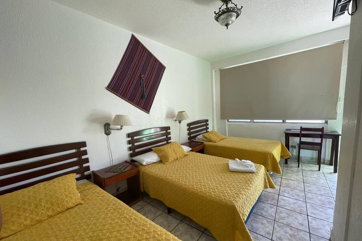A hotel room with three beds, each with a wooden headboard and yellow bedding. There are bedside tables with lamps, a window with a view, and a small table with a chair.