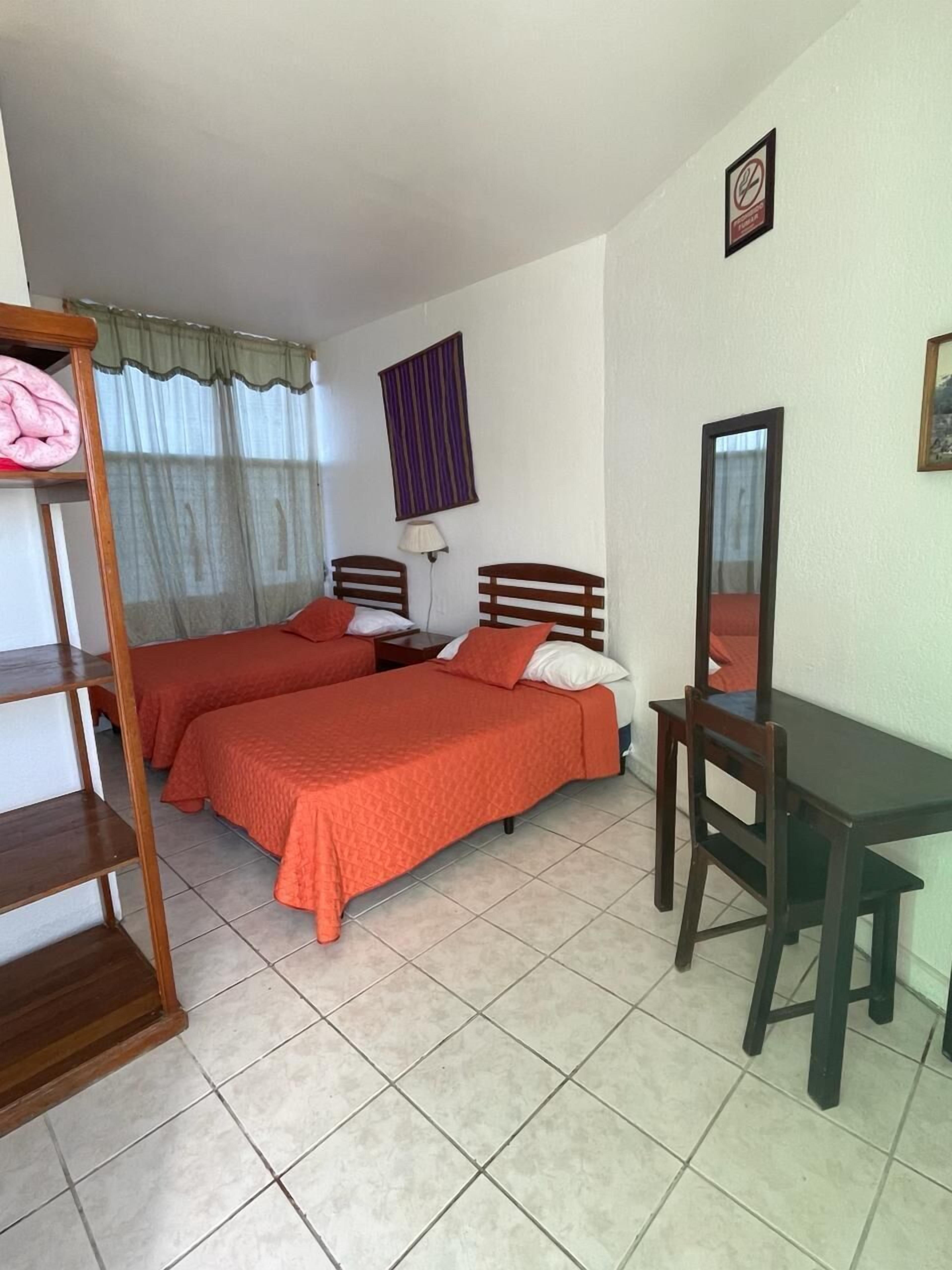 Una habitación con dos camas, un escritorio y una silla. Cuenta con una ventana con cortinas y una lámpara de pared.