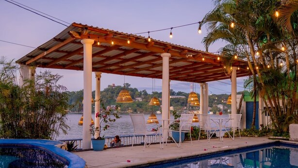 Un área junto a la piscina con una pérgola, sillas y luces colgantes.