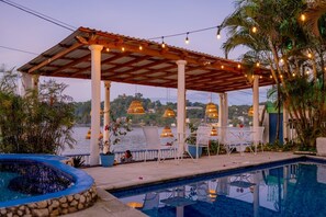 Un área junto a la piscina con una pérgola, sillas y luces colgantes.
