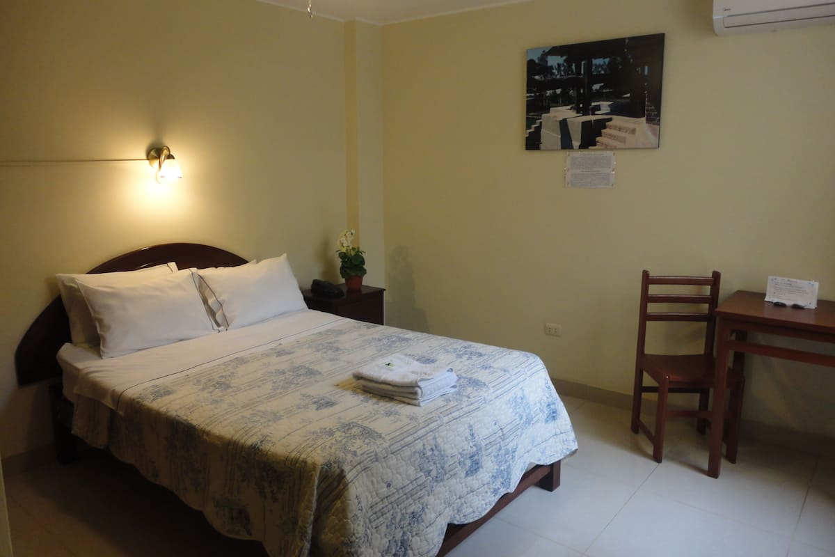 A neatly made bed with a patterned bedspread, a bedside table with a lamp, a chair, and a wall-mounted air conditioner.