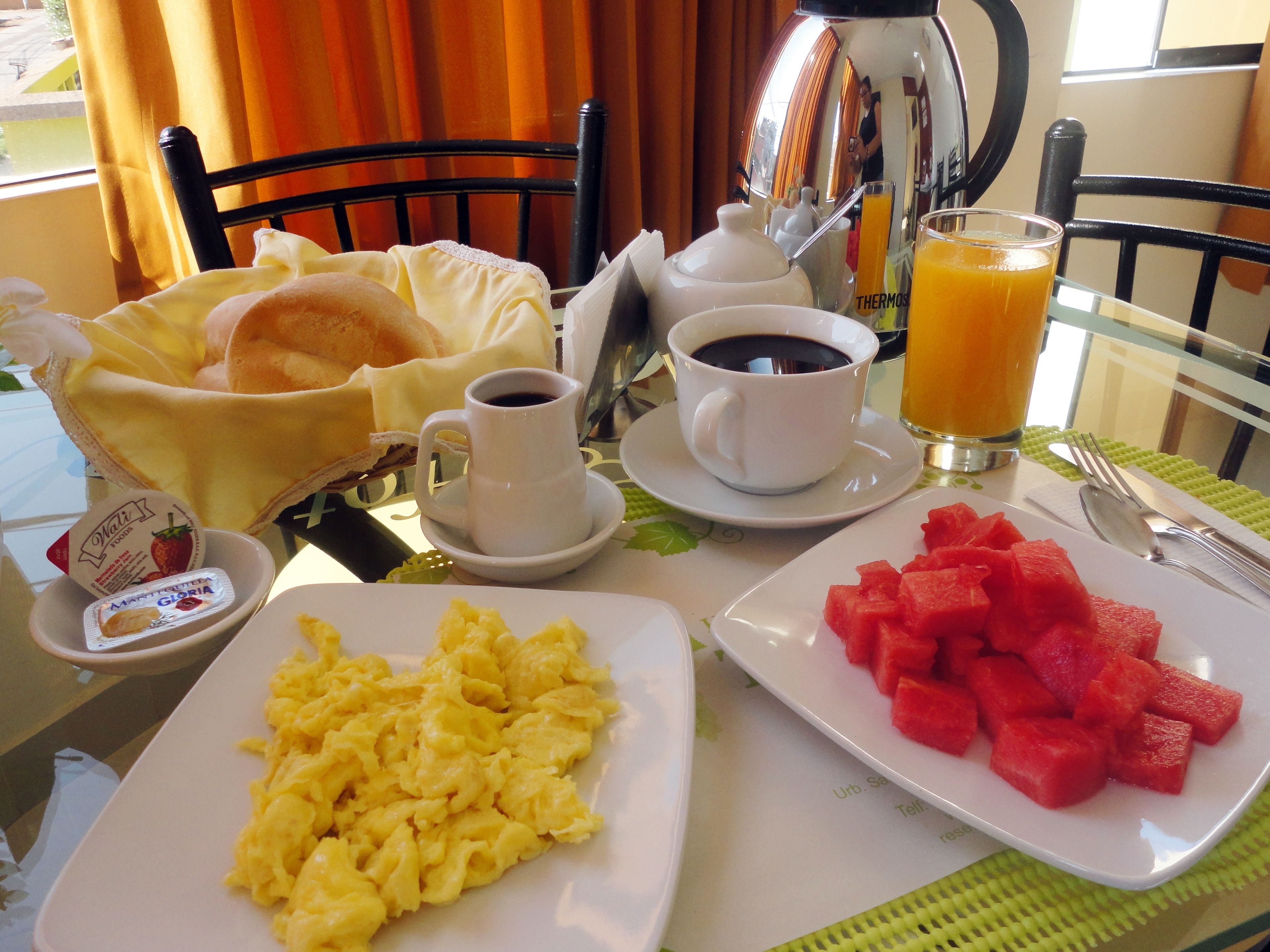 Una mesa de desayuno con huevos revueltos, sandía, café y jugo.
