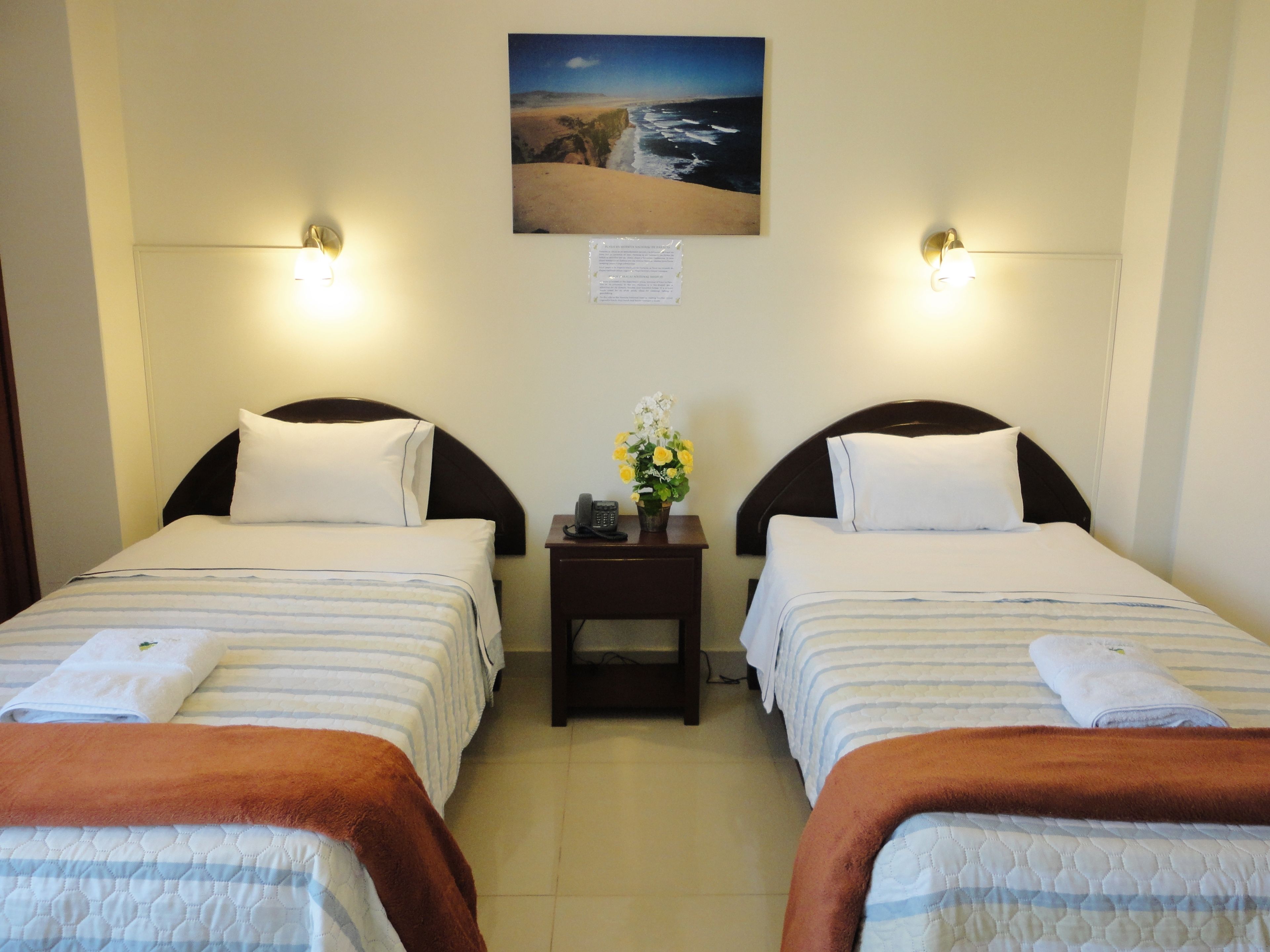 A hotel room with two beds, a nightstand with a phone and a vase of flowers, and a painting of a beach scene on the wall.
