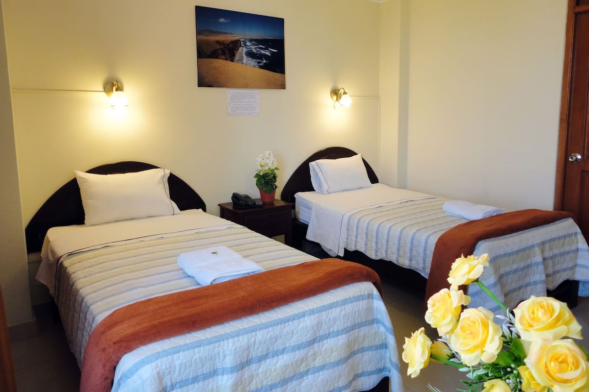 A hotel room with two beds, a nightstand with a phone, a painting of a beach scene, and a vase with yellow flowers.