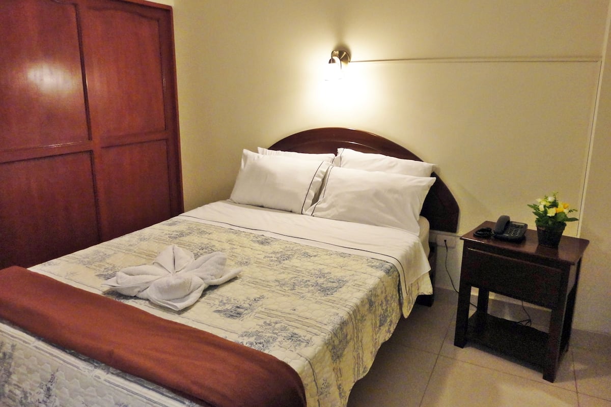 A neatly made bed with white linens and a folded towel, a bedside table with a phone and a small plant, and a wooden headboard.