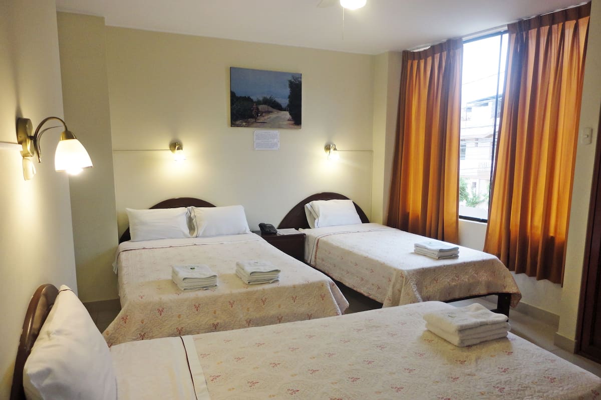 A hotel room with two single beds, each with a floral bedspread and a folded towel on the bed. There are two wall-mounted lamps, a painting on the wall, and a window with curtains.