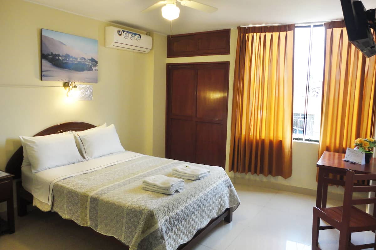 A hotel room with a bed, a desk, a chair, a ceiling fan, a window with curtains, and a wall-mounted TV.