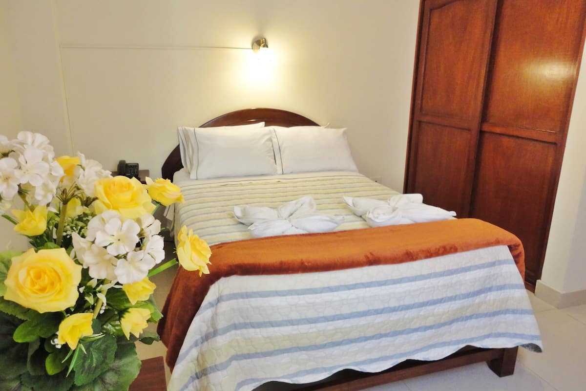A neatly made bed with a striped blanket and pillows, a wooden wardrobe, and a bouquet of yellow and white flowers on the side.