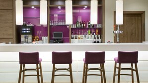 A bar area with three purple barstools, a coffee machine, and a variety of bottles and glasses on shelves. - Hampton by Hilton Oxford (Oxford)