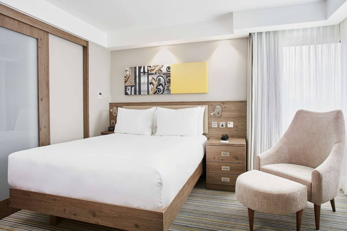 Habitación de hotel con una cama grande, una mesita de noche de madera, una silla y una ventana con cortinas.
