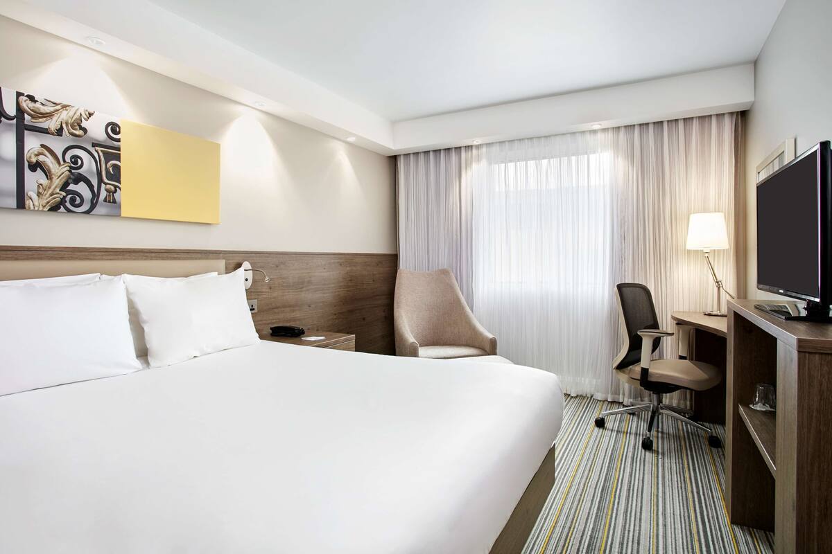Habitación de hotel con una cama grande, un escritorio con computadora, una silla, un televisor y un ventanal con cortinas.