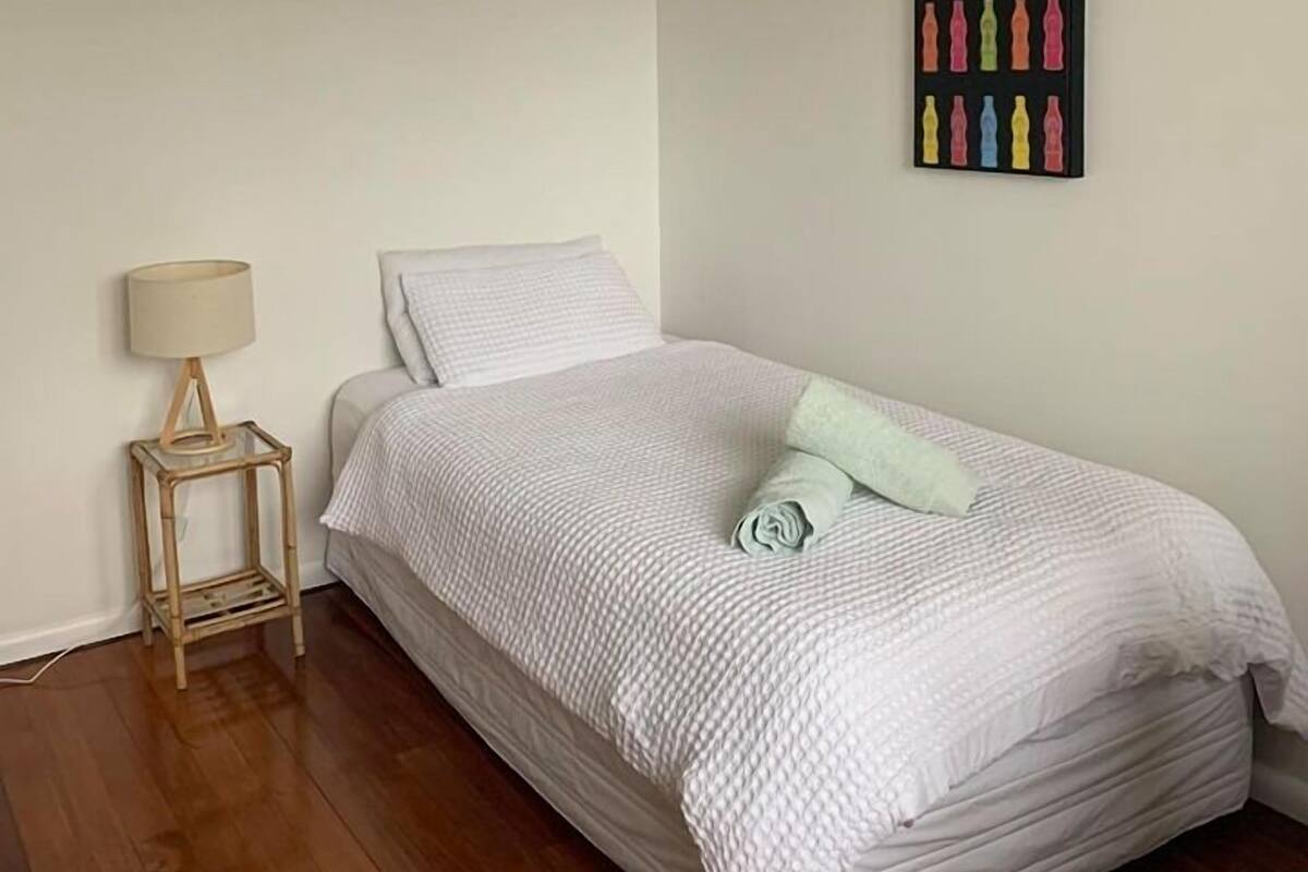 Ein Einzelbett mit weißer Daunendecke und grünem Kopfkissen, ein kleiner Nachttisch mit Lampe sowie ein Wandbild mit bunten Flaschen.