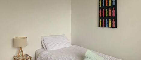 A single bed with a white duvet and a green pillow, a small bedside table with a lamp, and a wall art piece of colorful bottles.