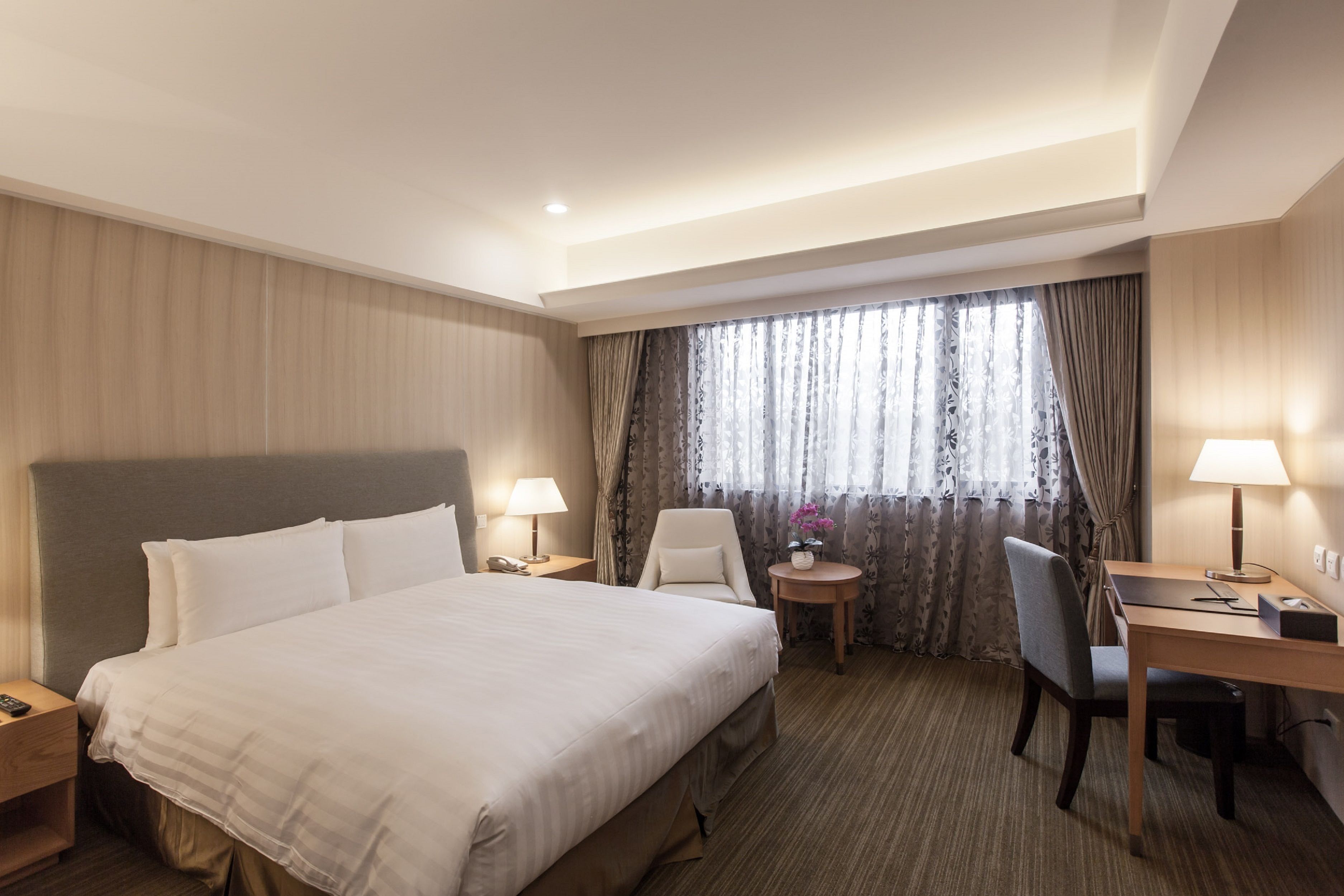 A hotel room with a large bed, a desk, a chair, a small table with a lamp, and a window with curtains.