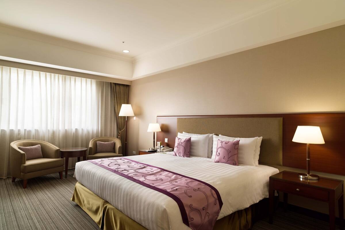 Une chambre d’hôtel avec un grand lit, deux tables de chevet, une chaise, une lampe et une fenêtre avec des rideaux.