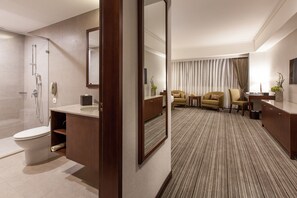 Une salle de bain moderne dans un hôtel, équipée d’une douche, de toilettes et d’un meuble-lavabo. On y trouve également un espace salon avec un canapé et des fauteuils.