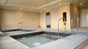 Une grande piscine intérieure à l’eau claire, entourée d’une terrasse en pierre et de rampes en métal.