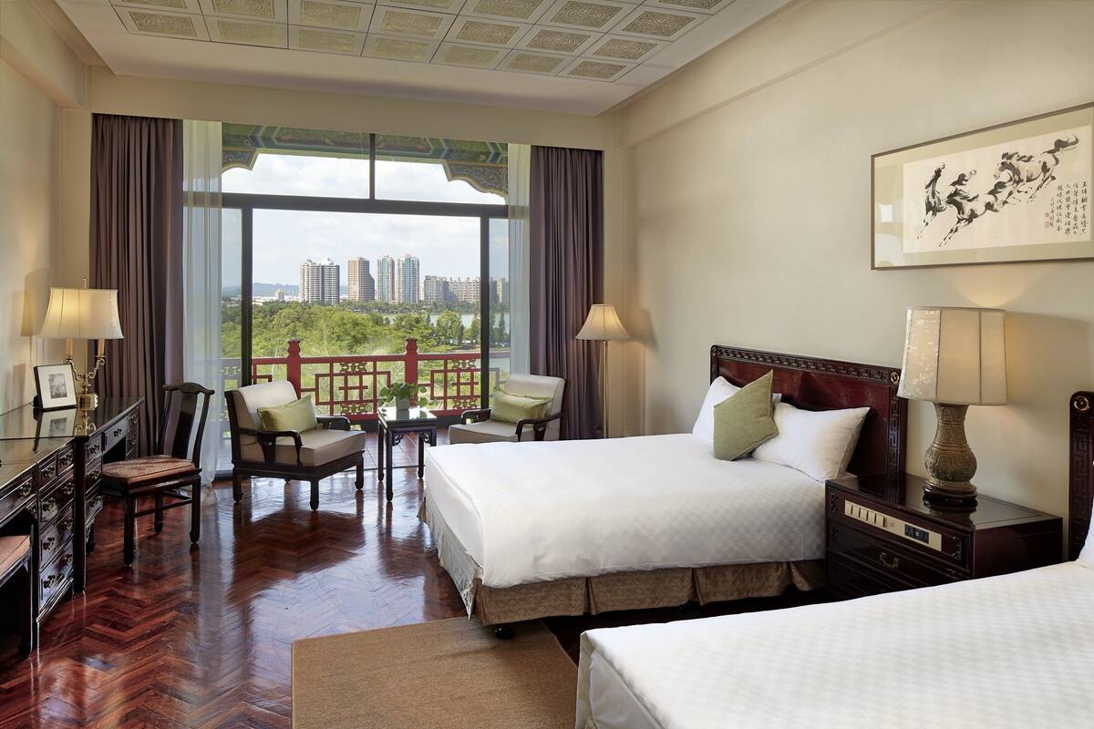 A hotel room with two beds, a desk, a chair, and a view of a cityscape through large windows.