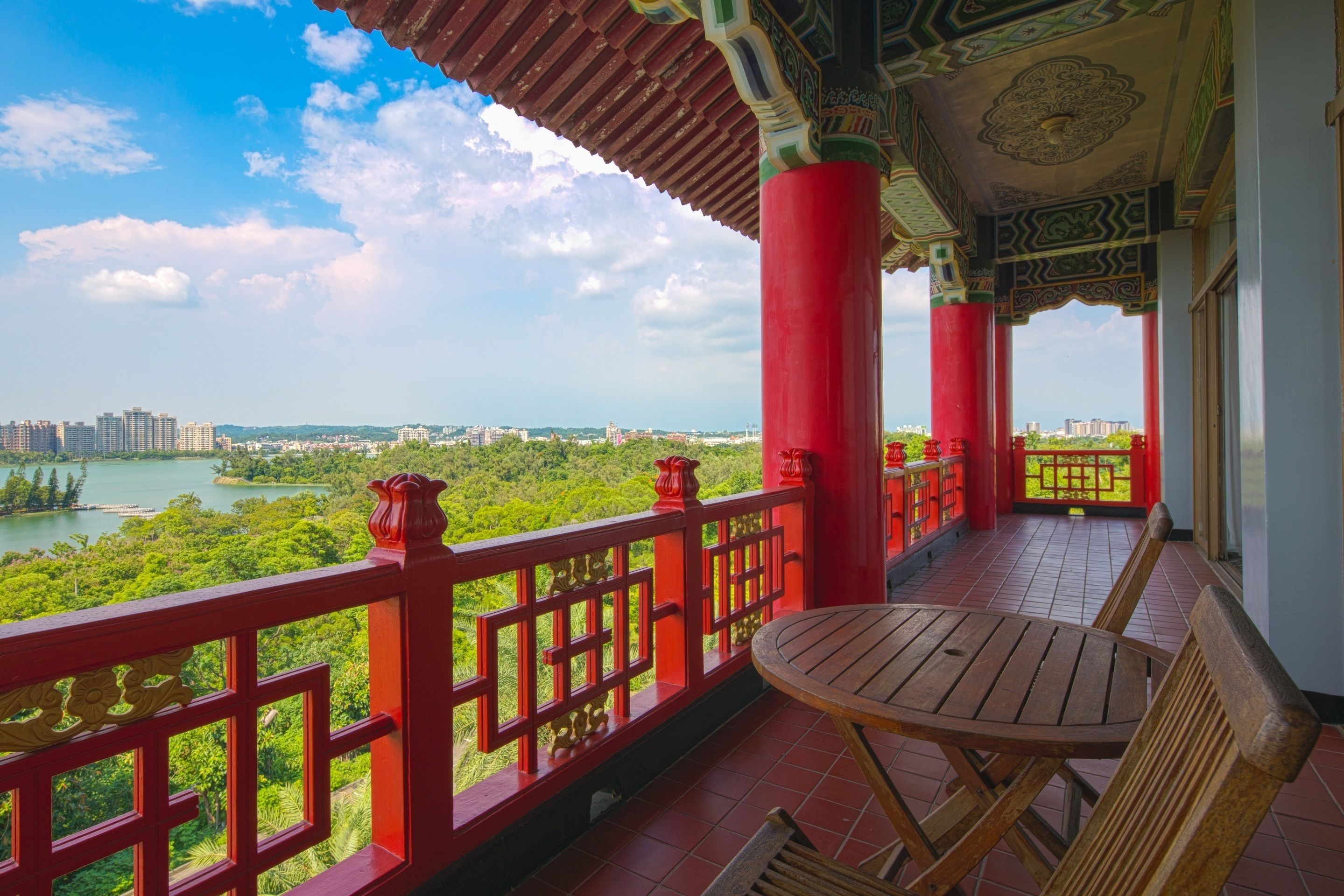 A balcony with a view of a lake and cityscape, featuring red railings and a wooden table.