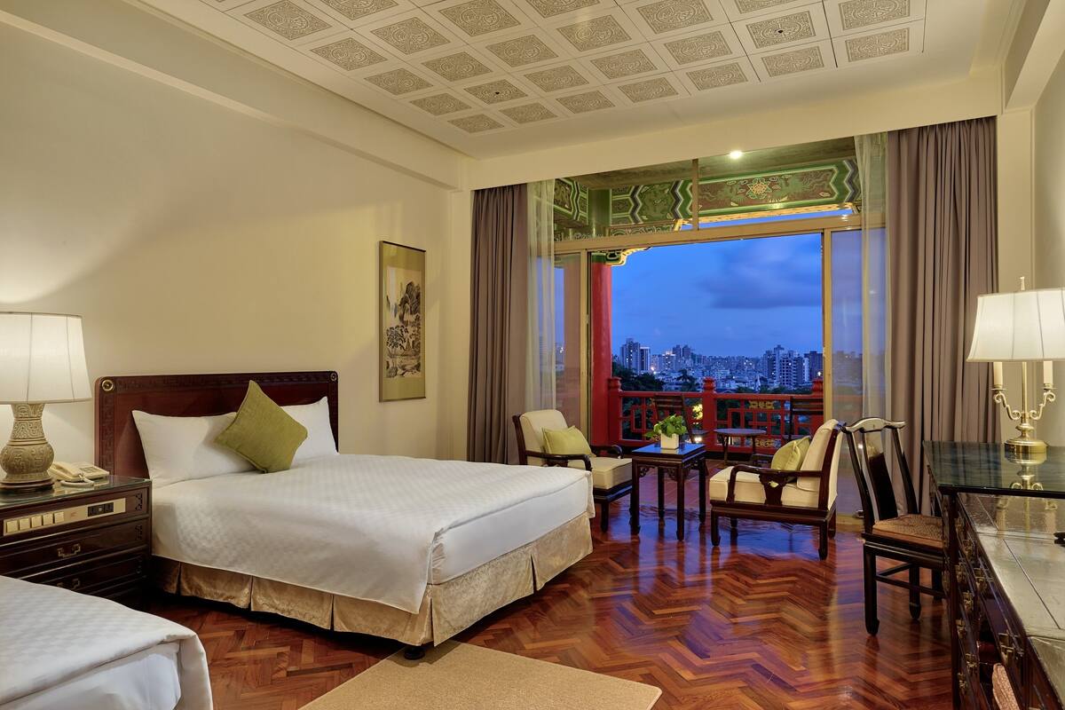 A hotel room with a large bed, a desk, two chairs, and a view of a cityscape at dusk.
