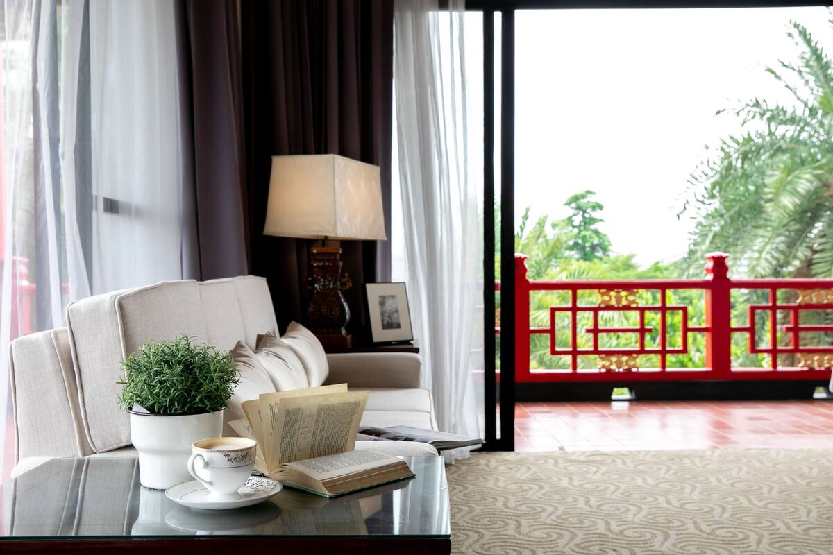 A room with a sofa, a table with a book and a cup, a potted plant, and a view of a red gate and greenery outside.