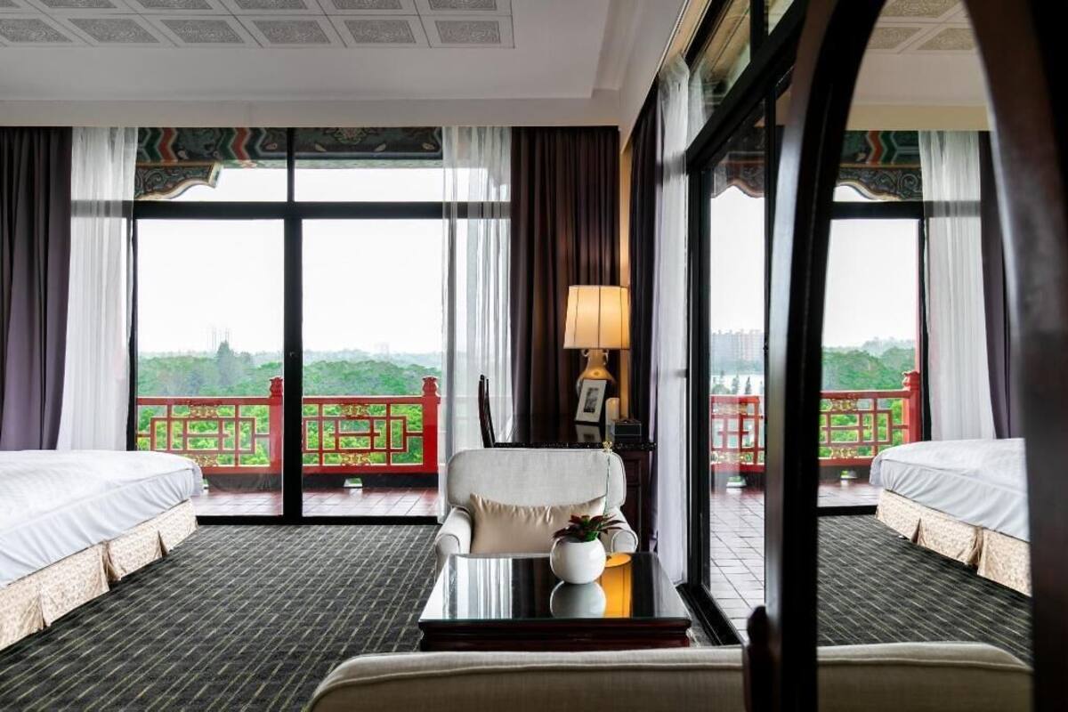 A hotel room with two beds, a sofa, a table with a vase, and large windows offering a view of a red balcony.