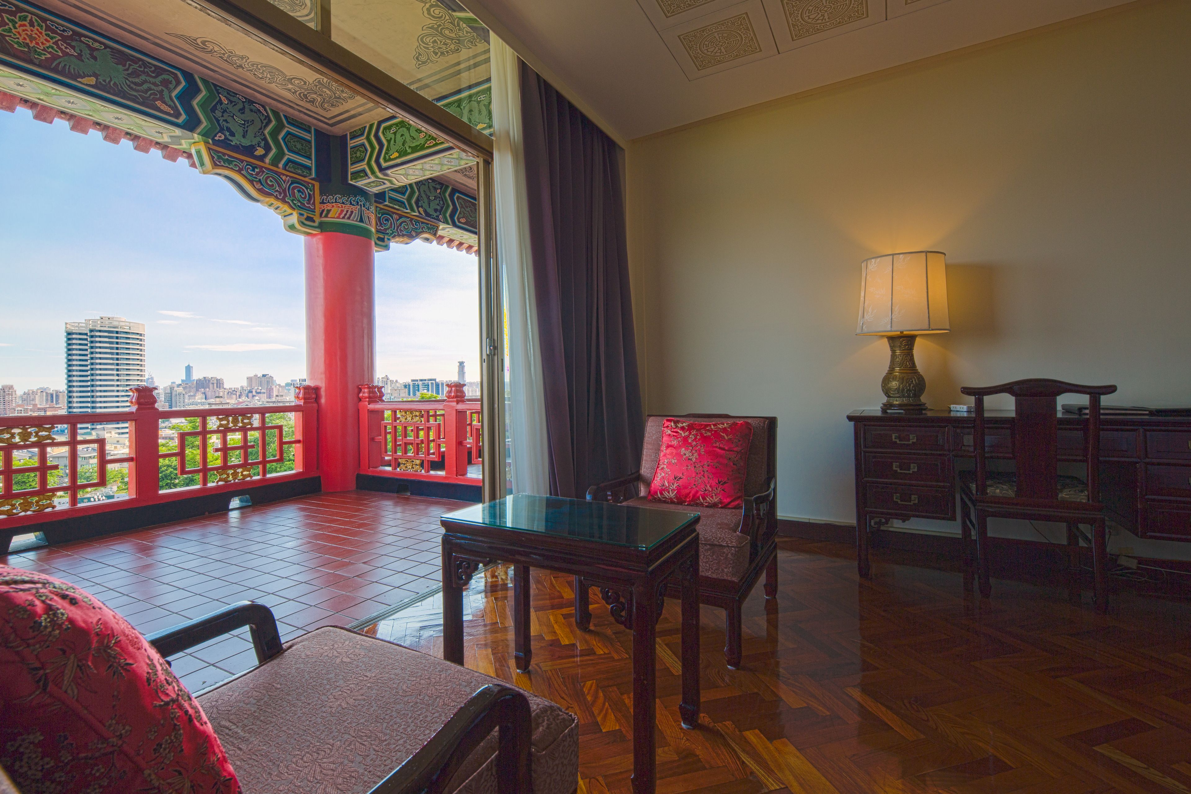 A room with a balcony, a table, a chair, a lamp, and a view of the cityscape.