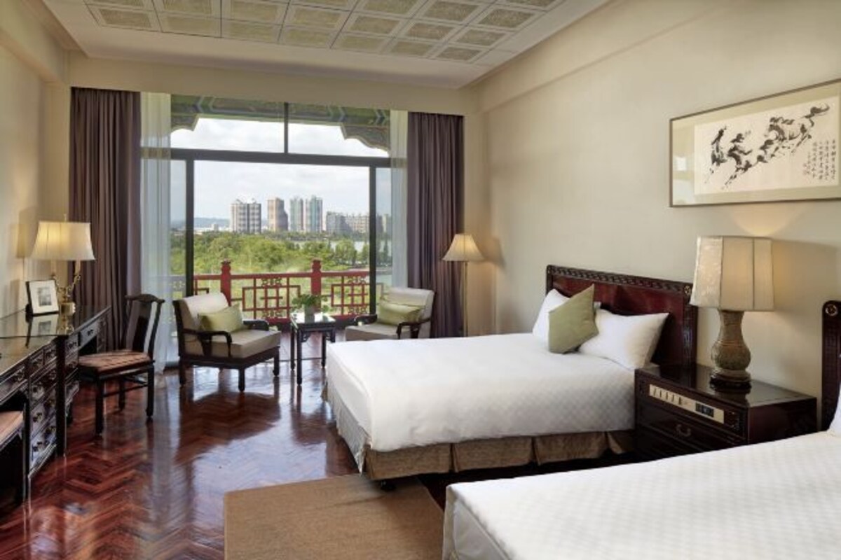 A hotel room with two beds, a desk with a chair, a small table with a chair, and a view of a cityscape through large windows.