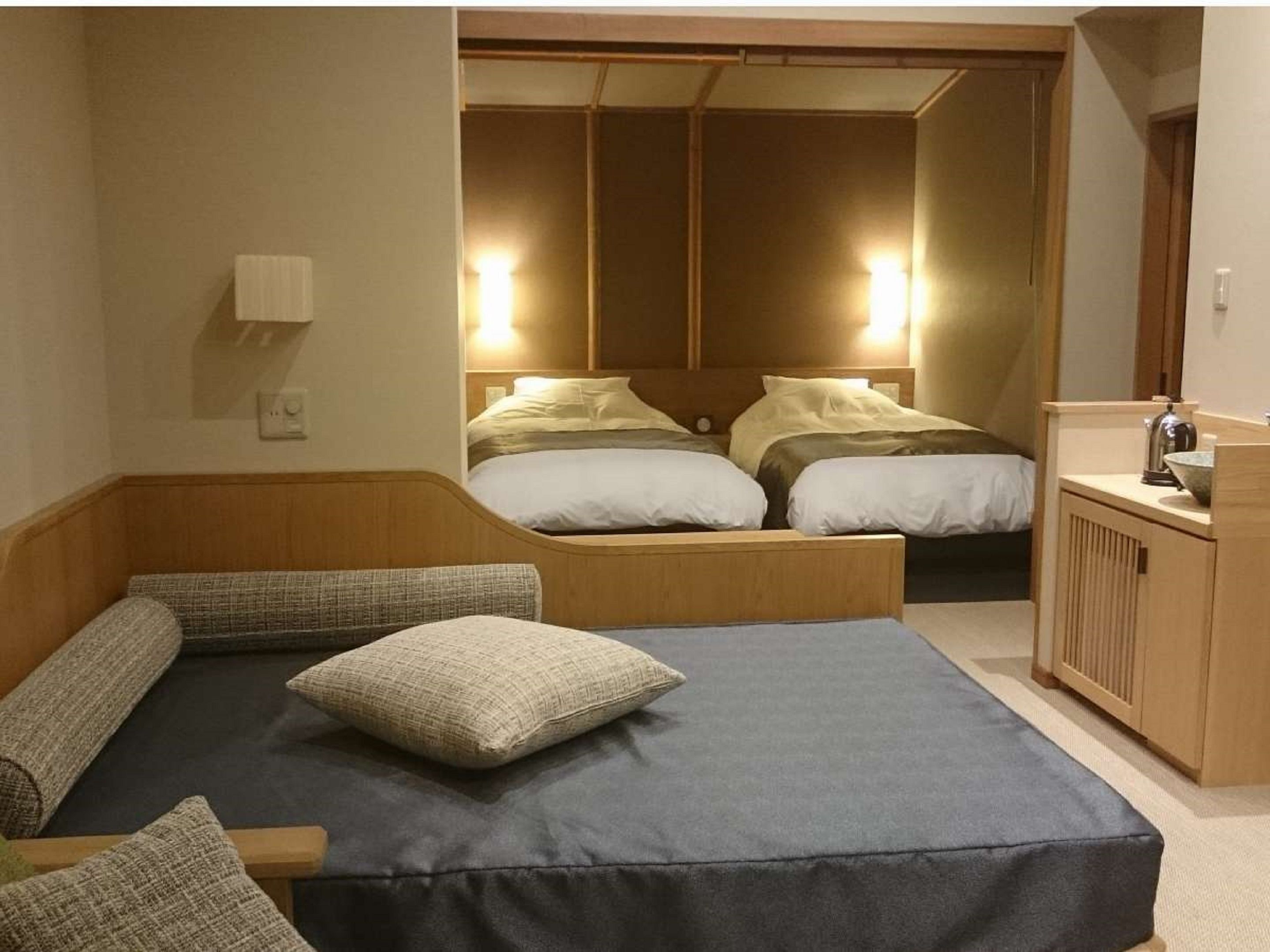 A hotel room with a large bed, a smaller bed, a desk with a kettle, and a cabinet.