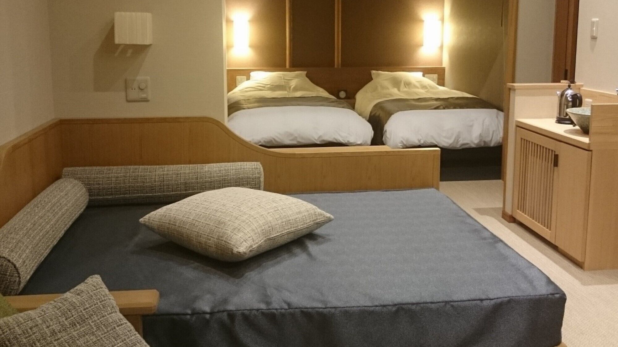 A hotel room with a large bed, a smaller bed, a wooden bench, and a desk with a kettle.