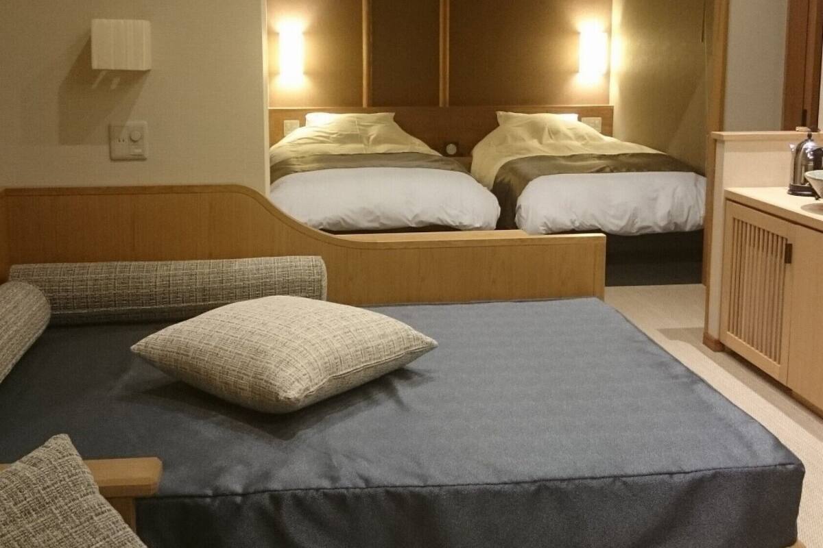 A hotel room with a large bed, a smaller bed, a wooden bench, and a desk with a kettle.
