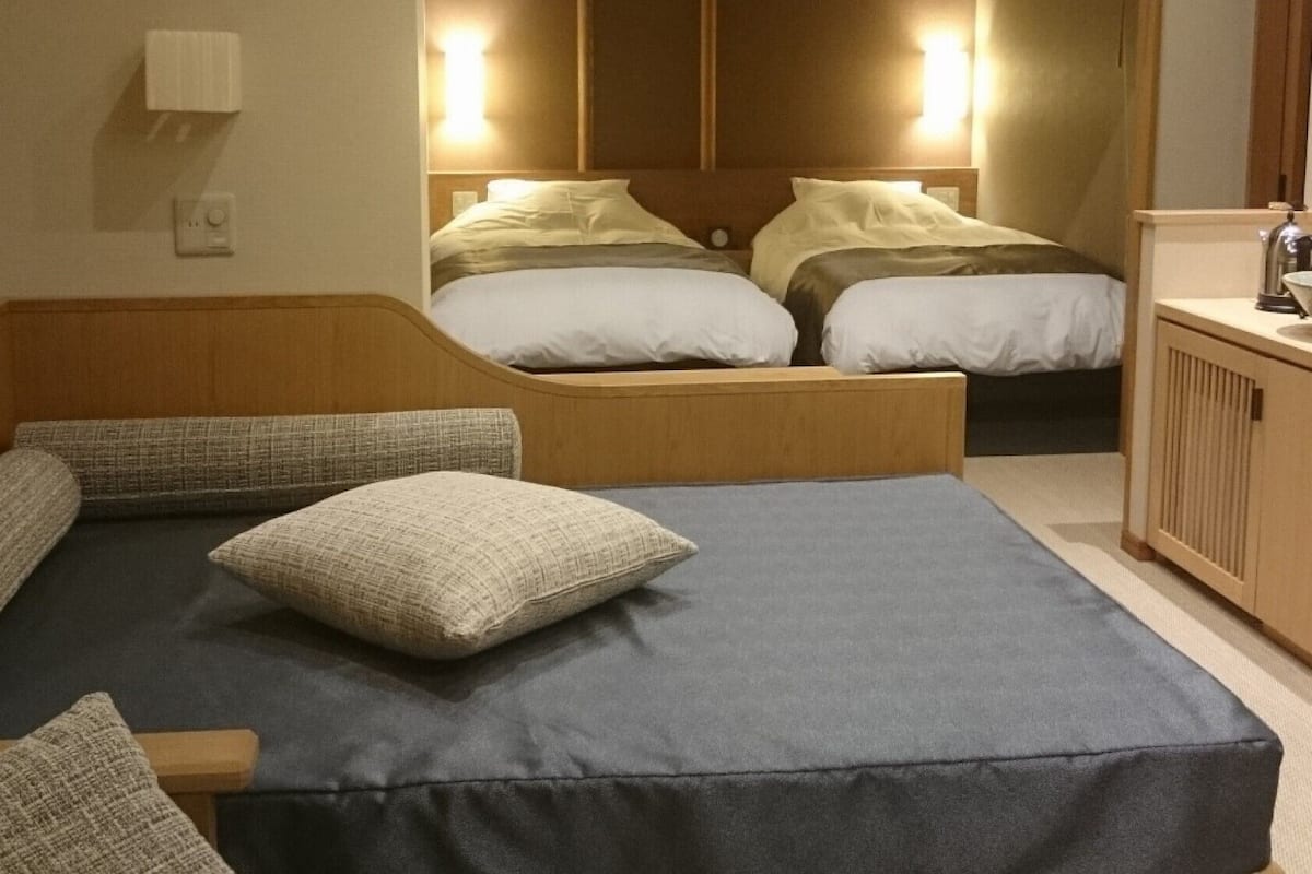 A hotel room with a large bed, a smaller bed, a wooden bench, and a desk with a kettle.